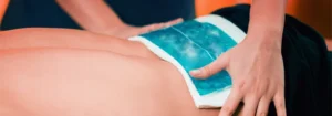 A blue gel cold pack wrapped in white cloth being applied to a person's lower back by a hand.
