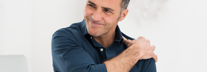 A man in a blue shirt holds his left shoulder with his right hand, looking away with a pained expression.