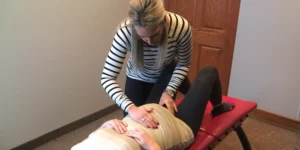 A practitioner in a striped shirt examines a patient lying on a red table with hands on her abdomen.