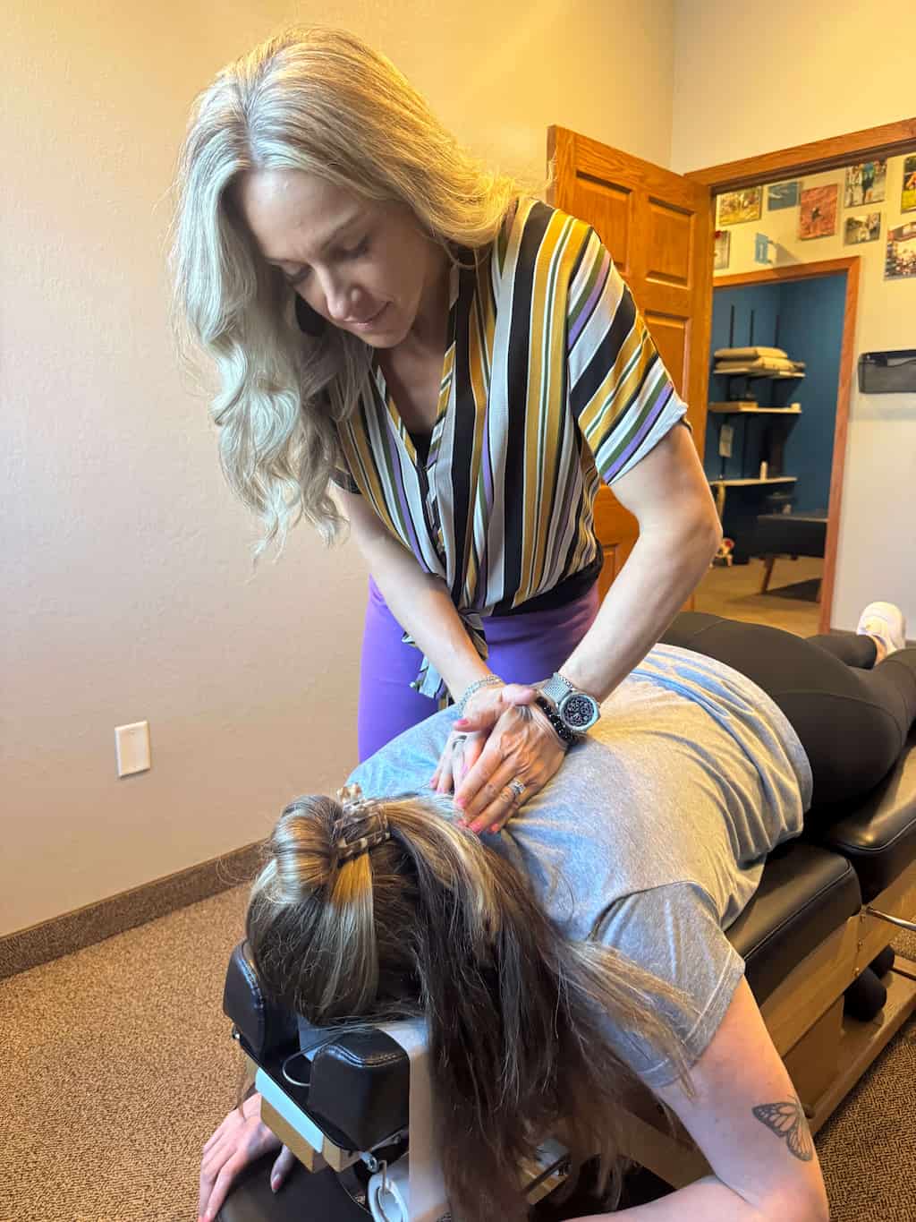 A chiropractor in a striped shirt adjusts a patient's upper back and neck on a chiropractic table.