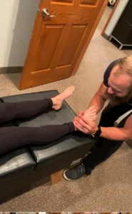 A practitioner with a beard manipulates a patient's bare foot and ankle on a black examination table.