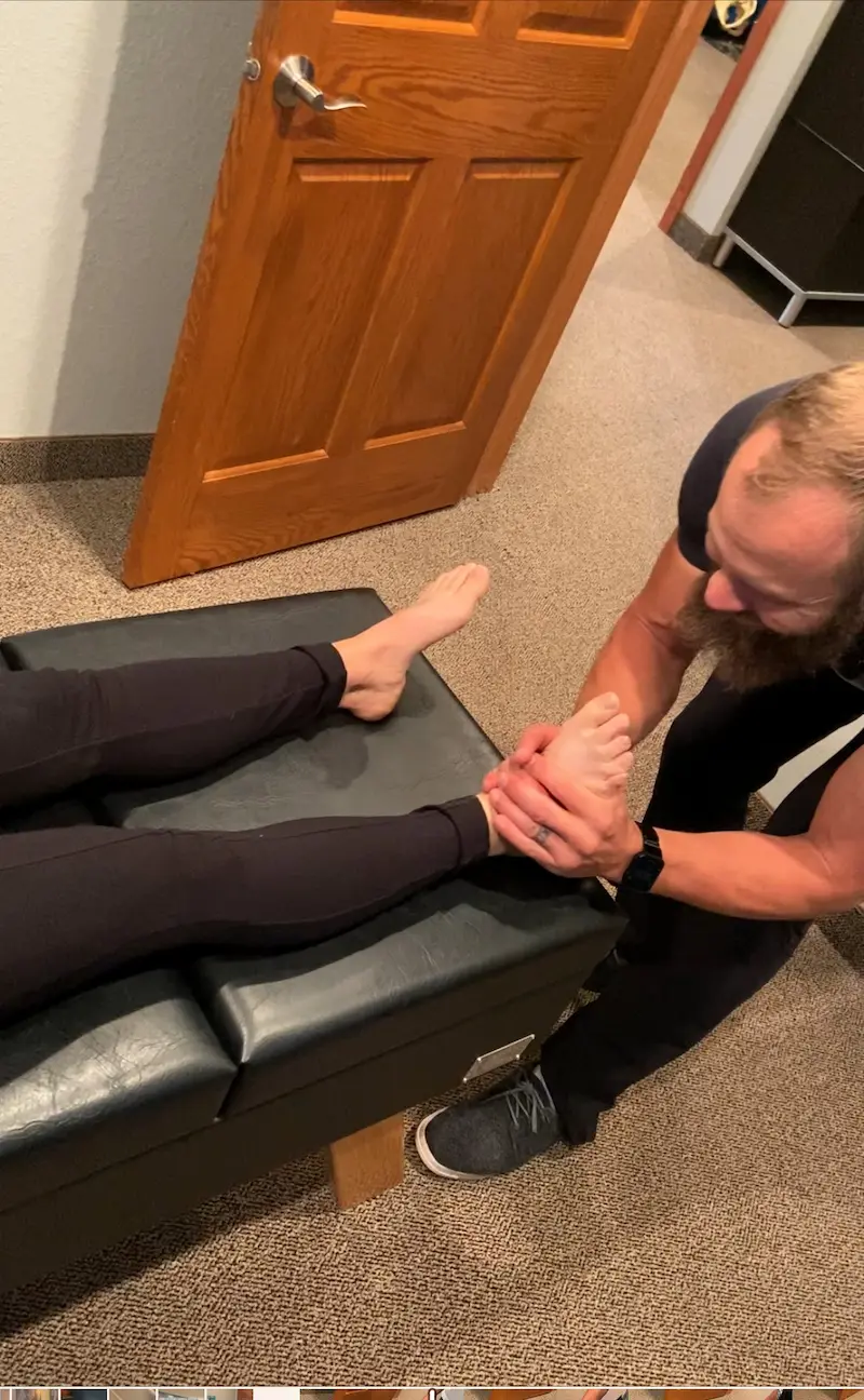 A practitioner with a beard manipulates a patient's bare foot and ankle on a black examination table.