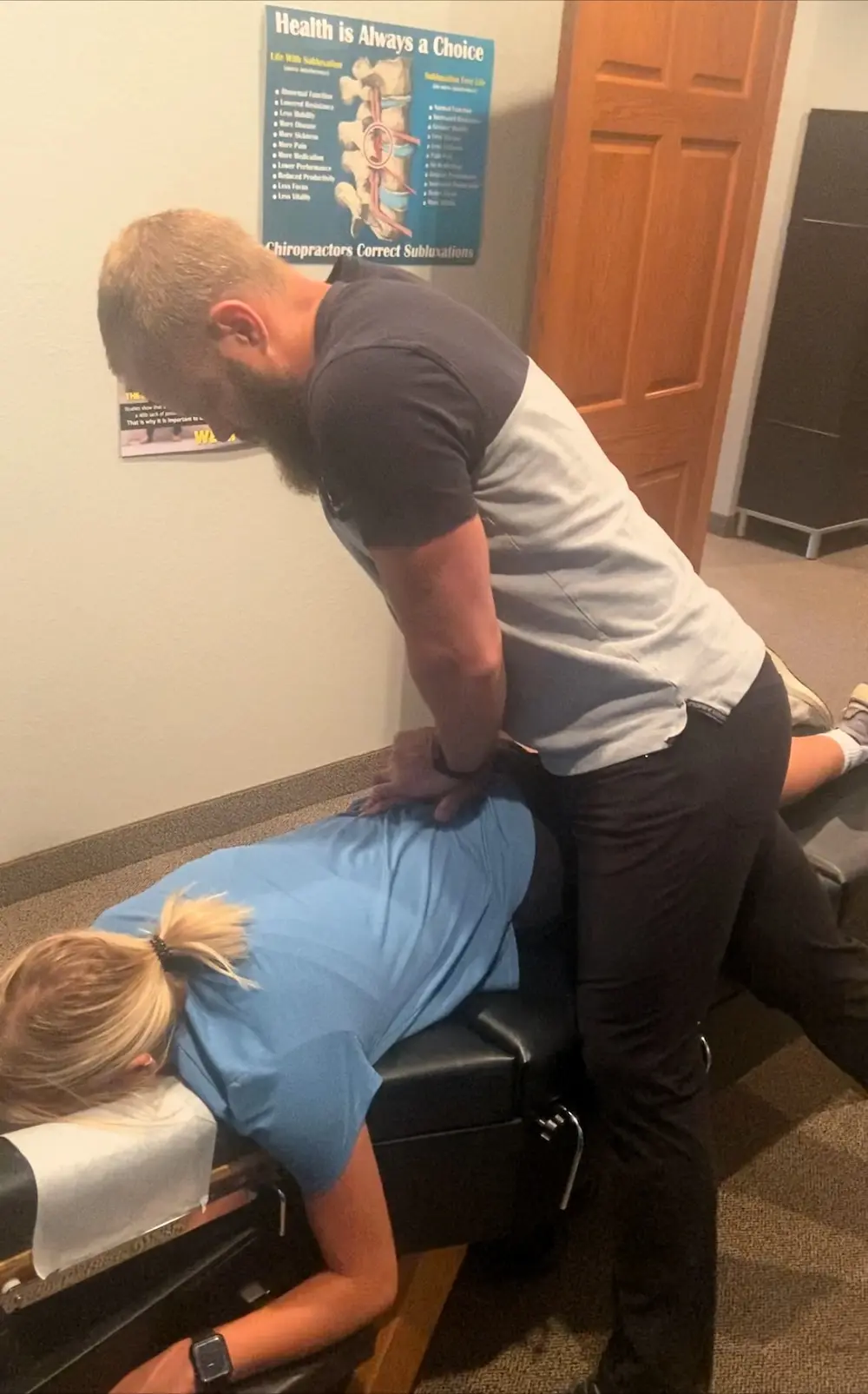 A male chiropractor adjusts a female patient's back as she lies face down on a chiropractic table.