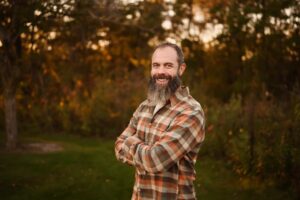 A smiling man with a dark beard and plaid shirt stands with crossed arms in an autumn outdoor setting.
