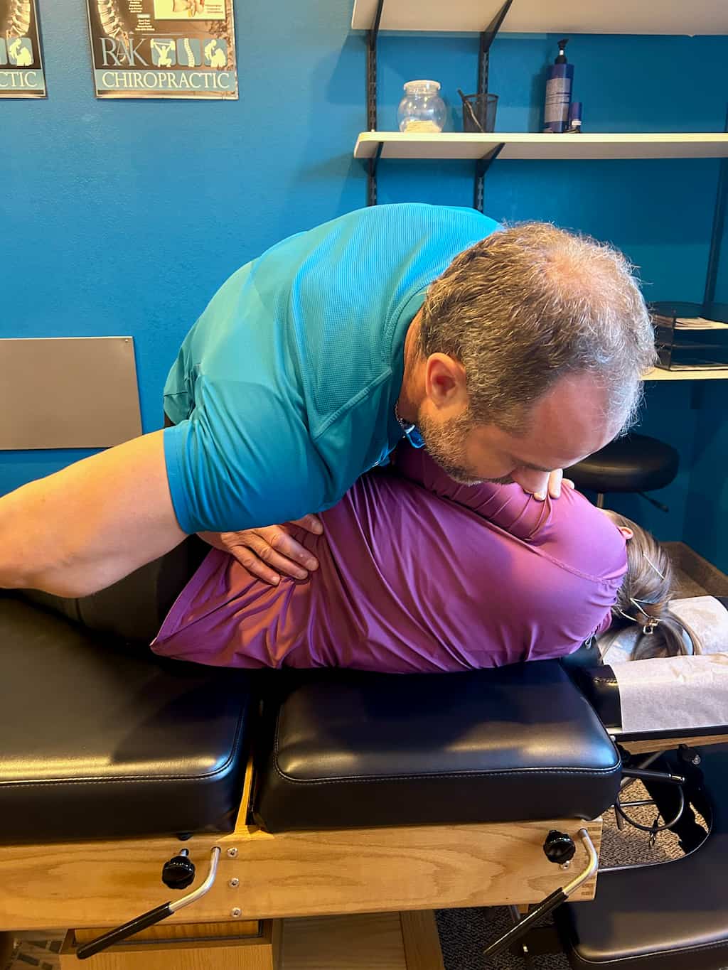 A male chiropractor adjusts a female patient's upper back on a chiropractic table.