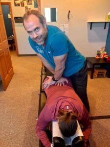 A male chiropractor adjusts the lower back of a female patient lying face down on a chiropractic table, smiling at the camera.