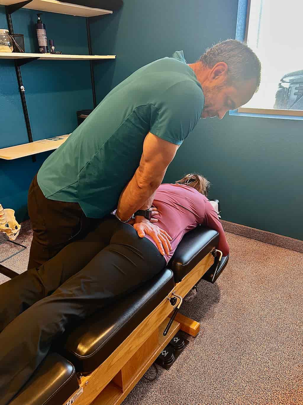 A chiropractor adjusts a woman's lower back as she lies face down on a black and wood chiropractic table.
