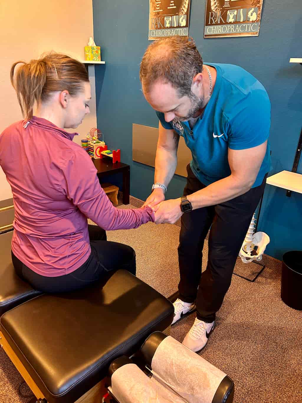 A chiropractor in a blue shirt adjusts a woman's wrist as she sits on a treatment table.
