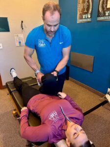 A male chiropractor adjusts a female patient's leg on a chiropractic table in an office setting.