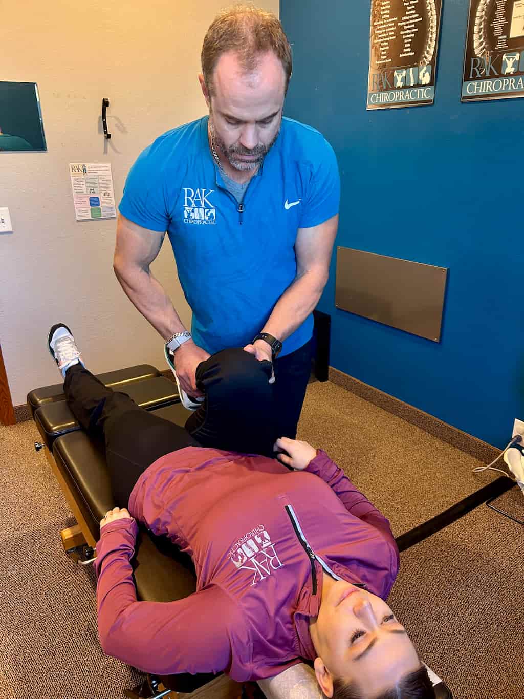 A male chiropractor adjusts a female patient's leg on a chiropractic table in an office setting.