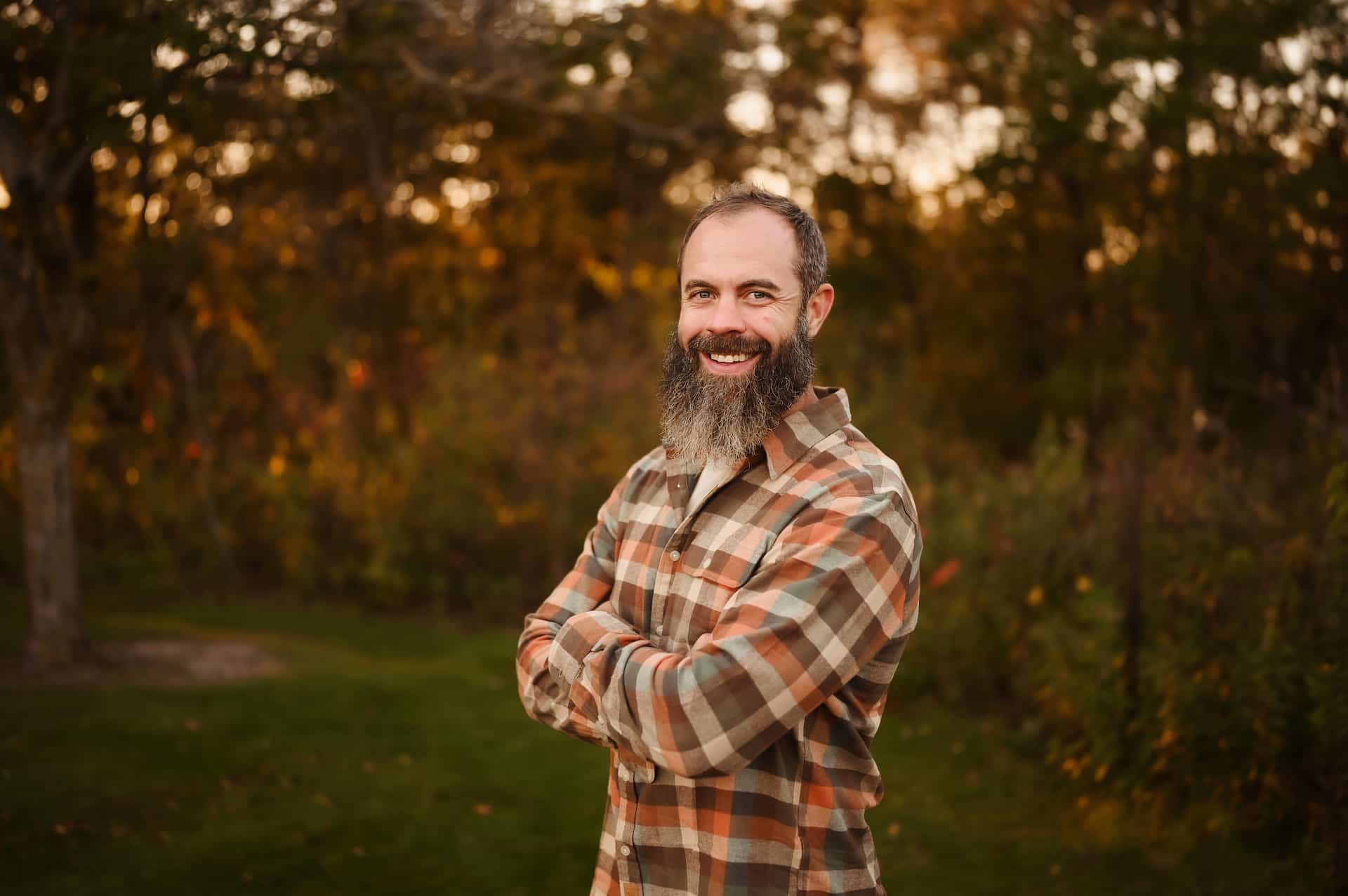 A smiling man with a dark beard and plaid shirt stands with crossed arms in an autumn outdoor setting.