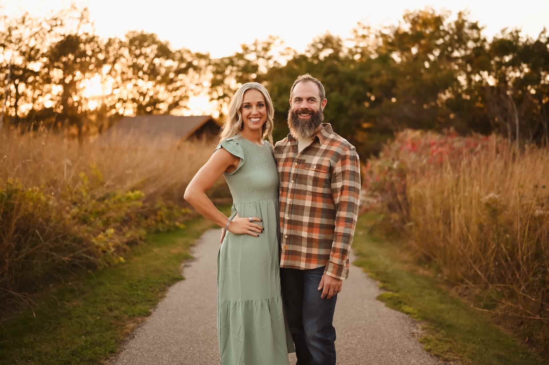 A smiling couple, a woman in a green dress and a man in a plaid shirt, stand on a path surrounded by autumn foliage at sunset.