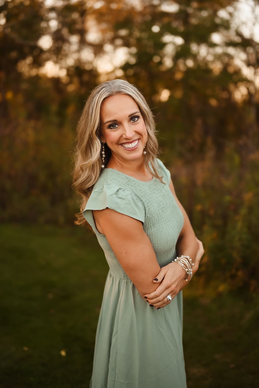 A smiling woman with long blonde and gray hair, wearing a sage green dress and pearl jewelry, stands with her arms crossed in an outdoor setting.