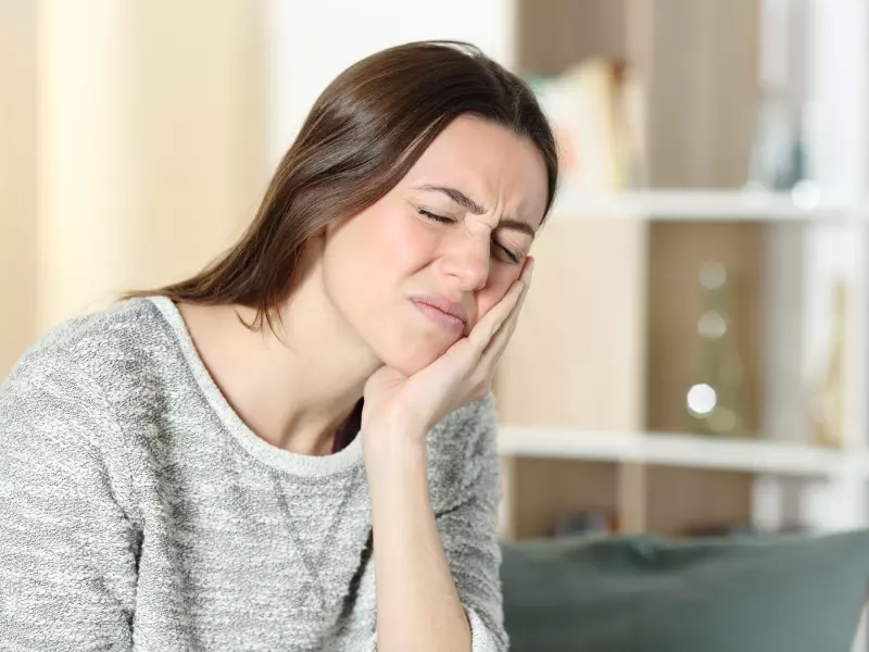 A young woman with closed eyes and a pained expression presses her hand against her cheek, indicating a toothache or jaw pain.