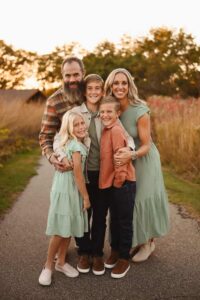 A family of five, two adults and three children, smiling on an outdoor path. The adults wear green and plaid, the children wear green and orange-brown.