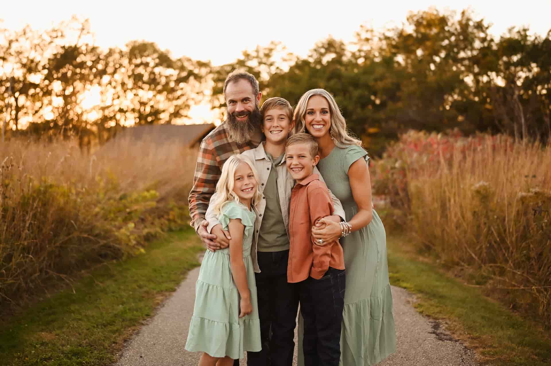 A smiling family of five, two adults and three children, stands on a path outdoors. The father has a beard and plaid shirt, the mother has blonde hair and a green dress. The three children, two boys and one girl, are also smiling. The background is a field with tall grass and trees at sunset.