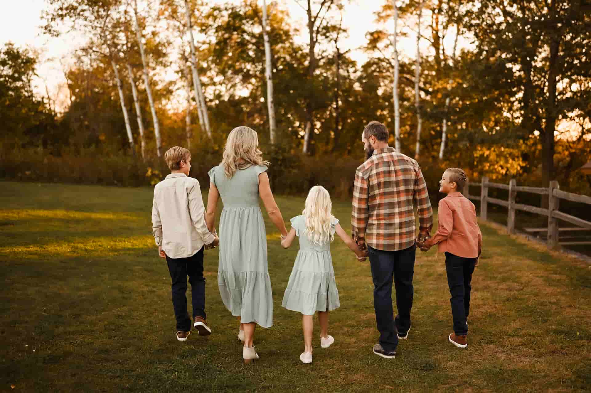 A family of five, two adults and three children, walk away from the camera holding hands in a grassy field with trees in the background during golden hour.