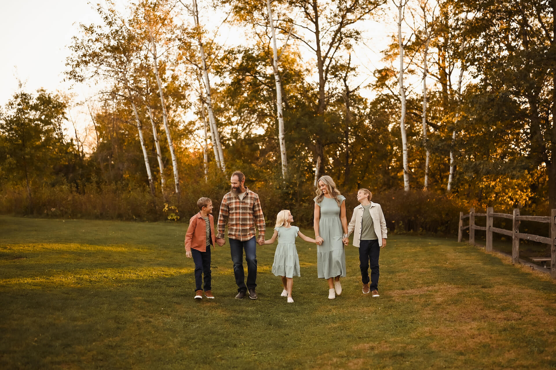 A family of five, two adults and three children, walks hand-in-hand across a grassy field with autumn trees in the background.