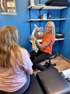 A blond chiropractor smiles while holding a spine model, explaining it to a patient in a chiropractic office.
