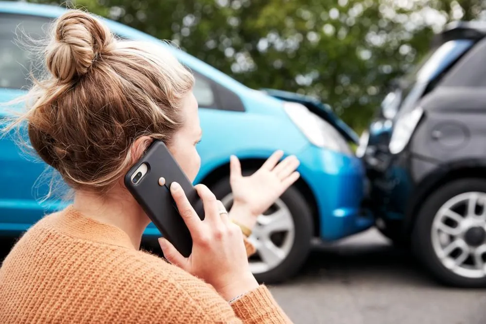 A woman with blonde hair on a phone call, standing next to two cars with damaged front ends after a collision.