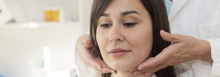 A medical professional's hands gently palpate the neck and jawline of a young woman with dark hair.