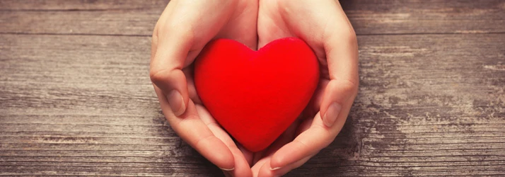 Cupped hands holding a red heart on a wooden background.