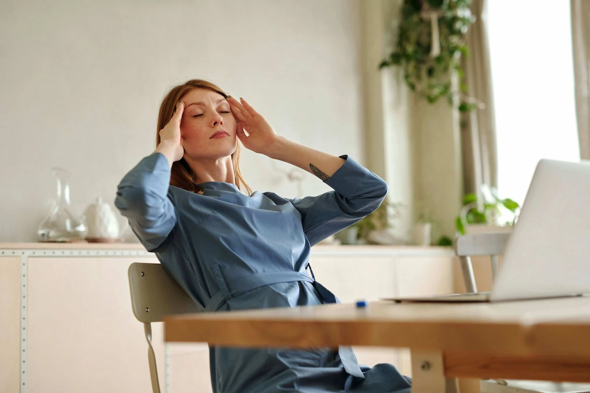 A woman with red hair and freckles sits at a table, eyes closed, pressing her hands to her temples as if experiencing a headache. A laptop is visible on the table.