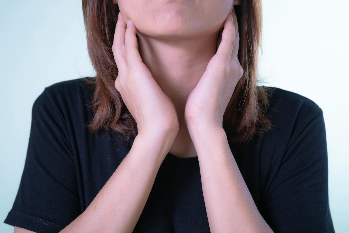 A person with light brown hair and a black t-shirt holds their hands to their neck, suggesting a sore throat or neck pain.