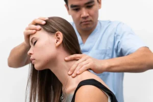 A man in blue scrubs adjusts a woman's neck and head. He holds her forehead with one hand and her neck with the other. The woman has long brown hair and closed eyes.