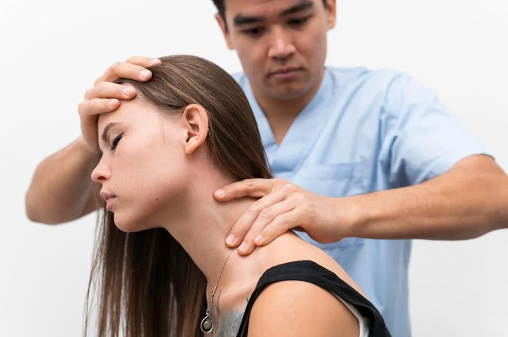 A man in blue scrubs adjusts a woman's neck and head. He holds her forehead with one hand and her neck with the other. The woman has long brown hair and closed eyes.