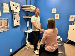 A smiling male chiropractor in a light green shirt shakes hands with a female patient sitting on a black adjustment table in a blue-walled office.