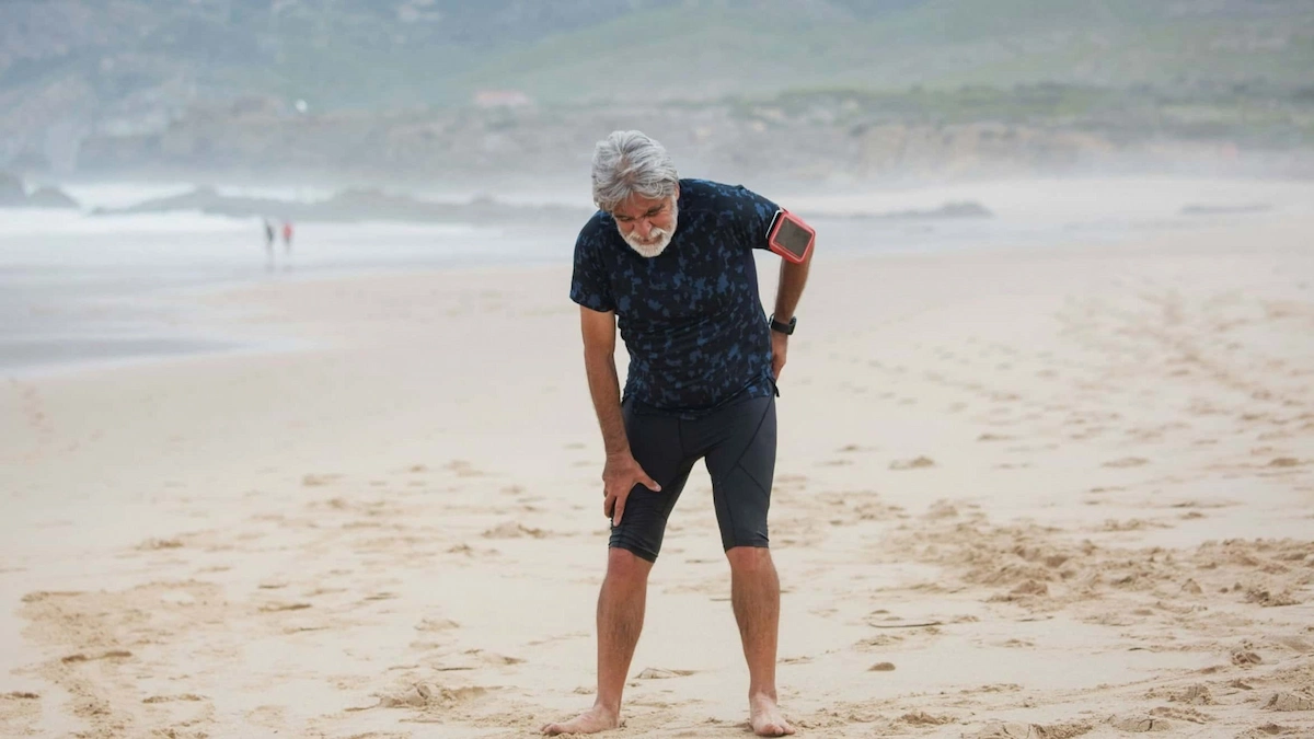 An older man with grey hair and a beard, wearing athletic clothes, stands barefoot on a sandy beach, bent over with hands on his knees, appearing to be in discomfort or stretching.