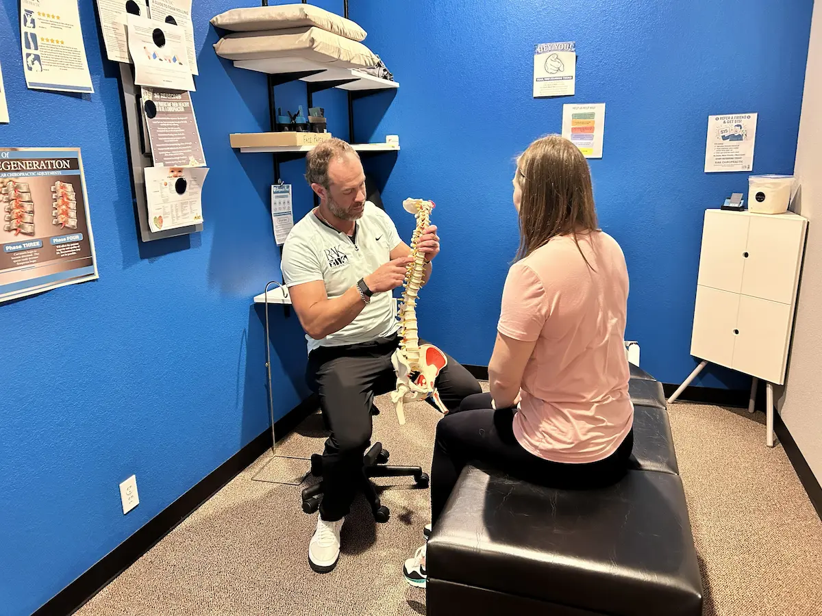 A chiropractor explains spinal anatomy using a skeletal model to a patient in a blue-walled consultation room.