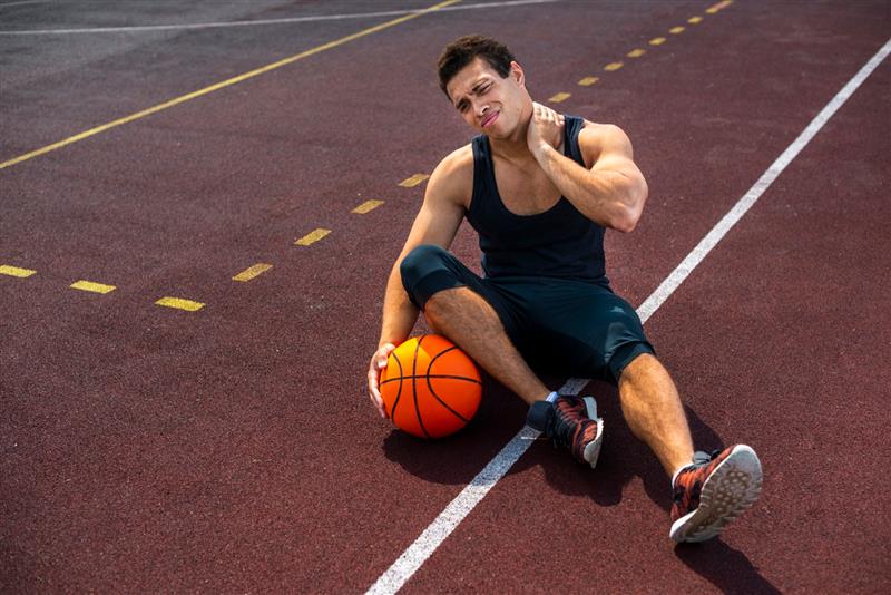 A male basketball player sits on a court, holding his neck in pain.