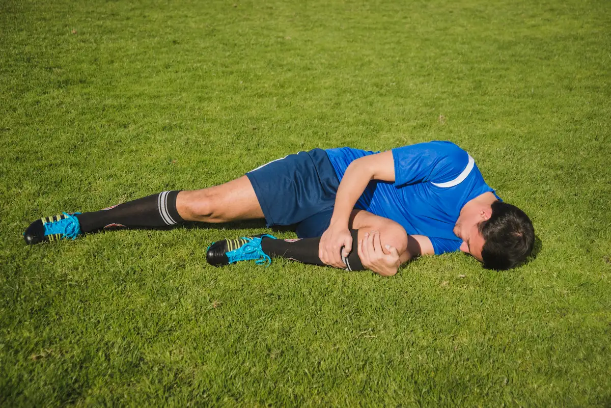 A male soccer player in a blue uniform lies on a green grass field, holding his knee with both hands, indicating an injury.