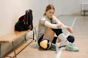 A female volleyball player sits on a gym floor next to a volleyball, backpack, and water bottle, looking down.