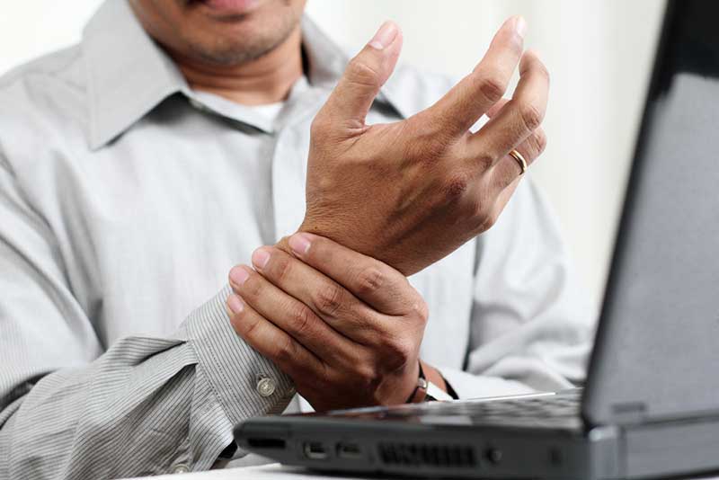 A person in a gray shirt holds their left wrist with their right hand, indicating pain, while sitting in front of a laptop.