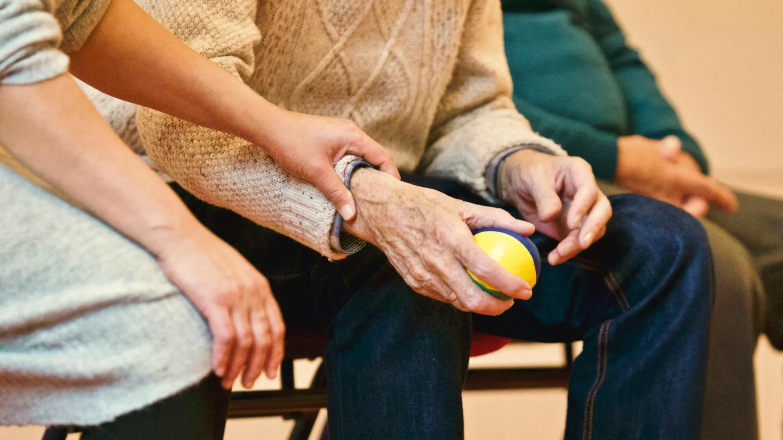 A younger person's hand rests on an older person's wrist, who holds a yellow, blue, and green ball.