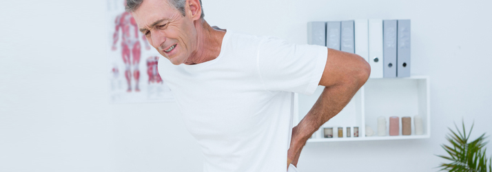 A man in a white t-shirt clutches his lower back, his face showing pain, in what appears to be a doctor's office with anatomical charts and shelves in the background.