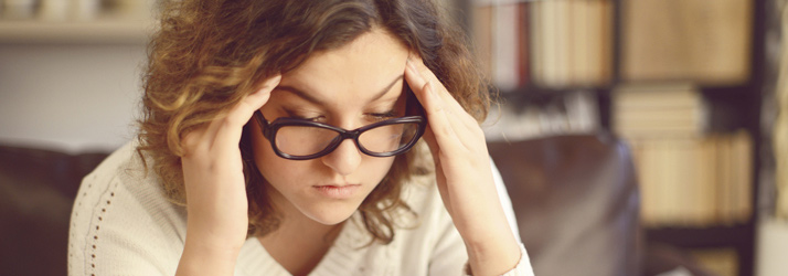 A young woman with curly hair and black glasses holds her temples, looking down with a pained or stressed expression.