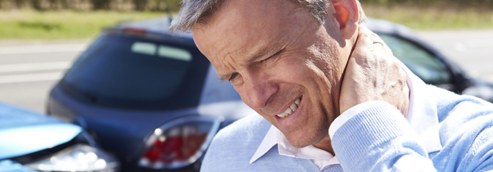 A man grimaces in pain, holding his neck, with two damaged cars visible in the blurred background, indicating a car accident.
