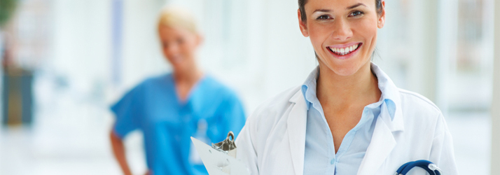 A smiling woman in a white lab coat with a stethoscope and clipboard, with a blurred person in blue scrubs in the background.