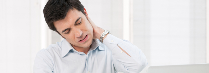 A man with dark hair, wearing a light blue shirt, holds the back of his neck with his left hand, eyes closed in discomfort, indicating neck pain.