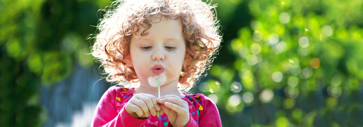 A young child with curly hair blowing on a dandelion, with seeds flying away, against a blurred green background.
