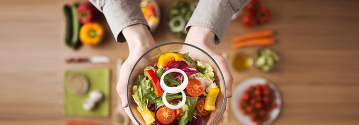 A person's hands hold a glass bowl of colorful salad with mixed greens, bell peppers, tomatoes, and onion rings, against a blurred background of various fresh vegetables on a wooden table.