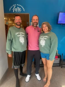 Three smiling people, two men and one woman, stand in front of a blue wall with "HEALING HAPPENS. here" written on it. Two wear green "Luke's Journey to Transplant" sweatshirts.